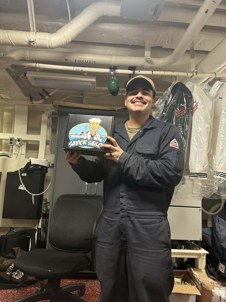 Sailor in coveralls holding a Savior Sailor care package aboard ship