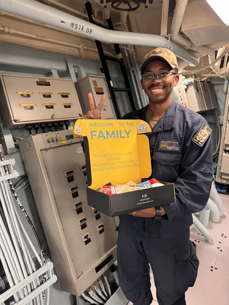 Sailor flashing a peace sign with an open Savior Sailor care package filled with snacks