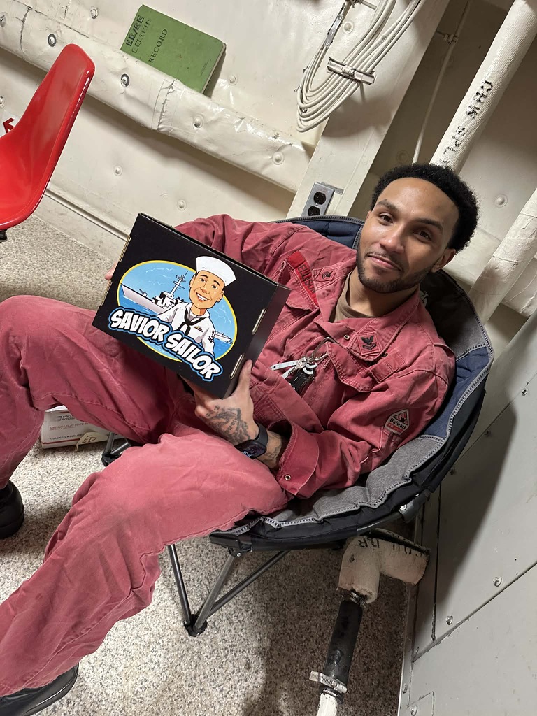 Sailor in red coveralls relaxing with his Savior Sailor care package aboard ship