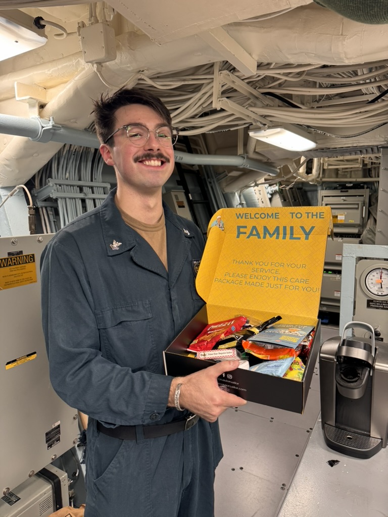Sailor with glasses showing an open Savior Sailor care package full of snacks