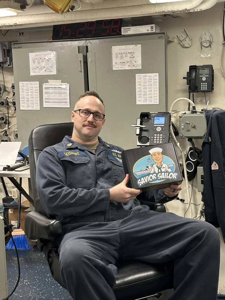 Sailor seated at a watch station holding his Savior Sailor care package